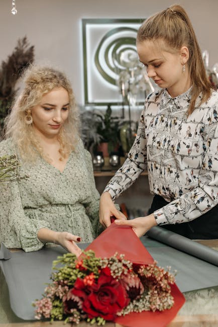 A woman with long, light brown hair styled in a ponytail, wearing a patterned blouse, is wrapping a floral bouquet with red and pink roses, baby's breath, and greenery in red wrapping paper on a workt