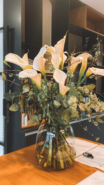 A large, elegant floral arrangement in a clear glass vase sits on a wooden table, featuring tall, creamy white calla lilies with smooth, trumpet-shaped petals and long, slender stems. Accompanying the