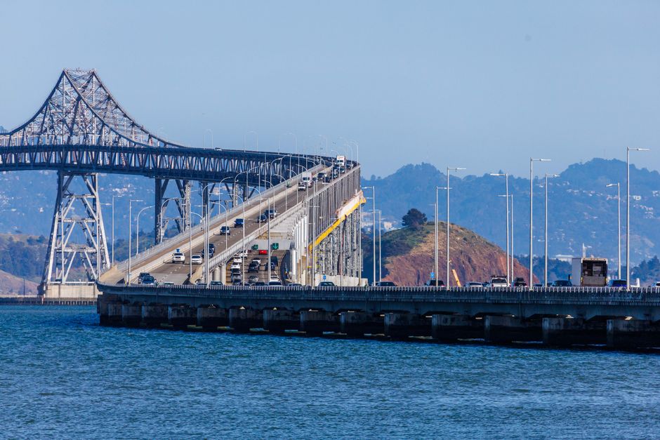 A view of the San Francisco-Oakland Bay Bridge extending over the water, with multiple lanes of traffic and a train traveling across the elevated structure. The bridge's steel framework and suspension