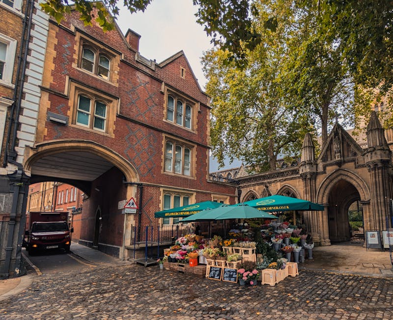 A vibrant flower stall positioned on a cobblestone street under a large teal umbrella, displaying an assortment of fresh flowers including roses, peonies, and tulips in various colors such as pink, ye