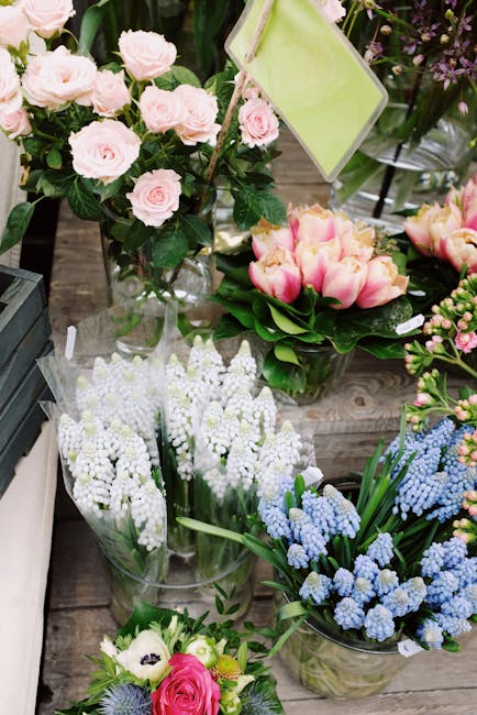 An arrangement of fresh flowers displayed on a wooden surface, featuring a bouquet of light pink roses with lush green leaves in a clear glass vase, a bunch of pale pink tulips with green stems and le