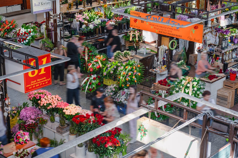 A bustling indoor flower market scene with a variety of fresh floral arrangements and bouquets displayed on white tables and stands. Prominent in the foreground are vibrant pink, red, and purple chrys