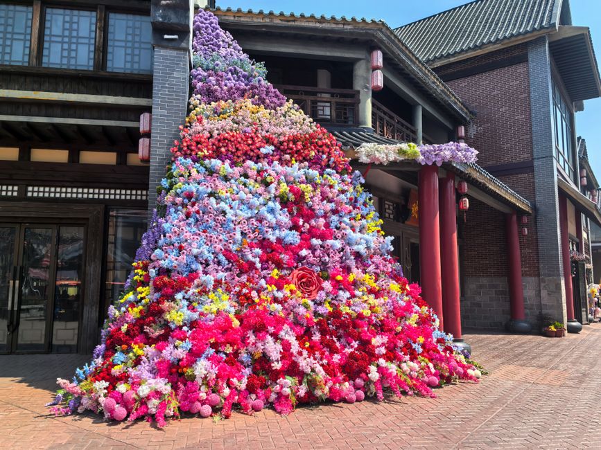 A large floral installation arranged outdoors in front of a traditional-style building with dark wooden beams, tiled roof, and red columns. The installation features a cascading pyramid of fresh flowe