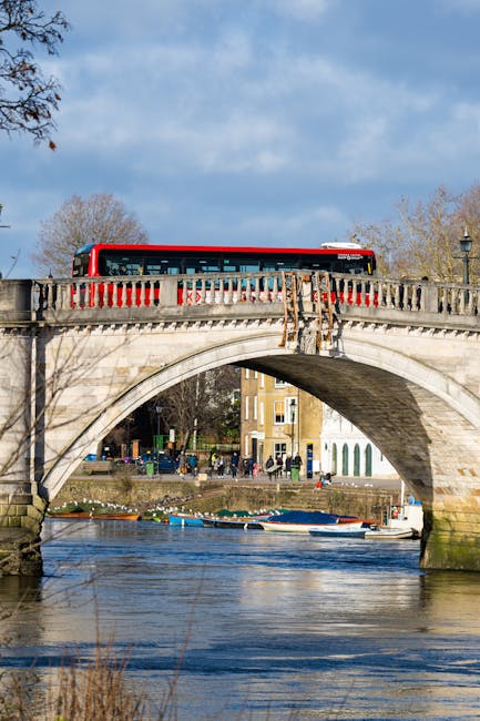 A red and white bus crossing a historic stone bridge over the River Thames in Putney, with a clear blue sky and scattered clouds overhead. Below the bridge, several small boats are moored along the ri