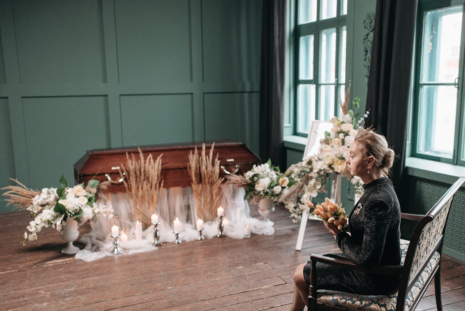 A woman seated in a vintage-style chair inside a softly lit room with green paneled walls and large windows, is holding a bouquet of peach-colored flowers. In front of her, a floral arrangement with w