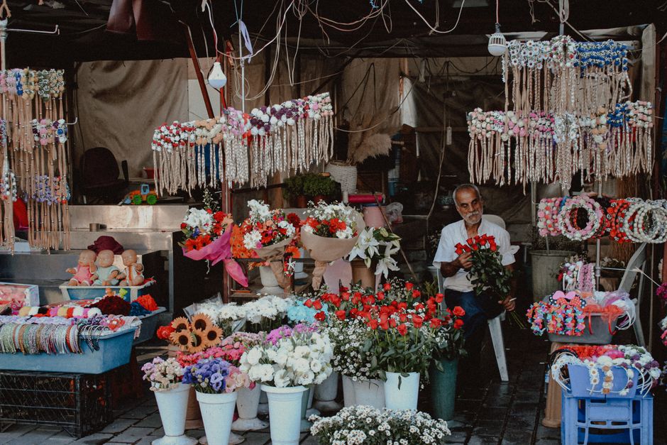 A flower stall with a variety of fresh flowers arranged in white and brown pots, including red roses, white daisies, purple and pink blossoms, and baby's breath. The flowers are displayed on tables an