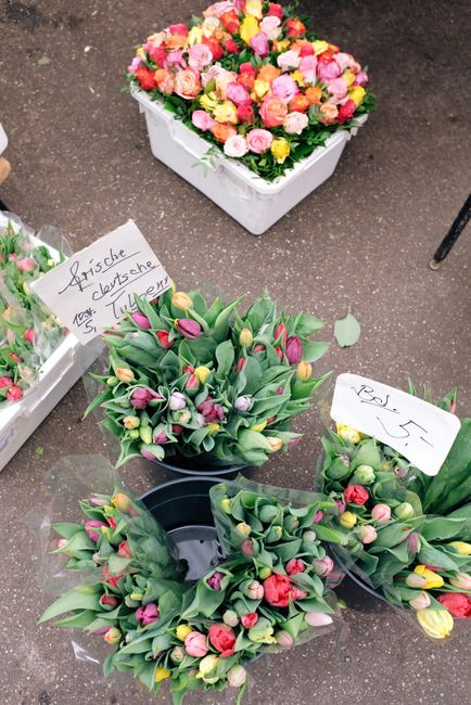 A vibrant assortment of fresh flower arrangements displayed on a marketplace floor, featuring a large, round bouquet of mixed pink, yellow, and peach roses in a white square box. In front, there are s