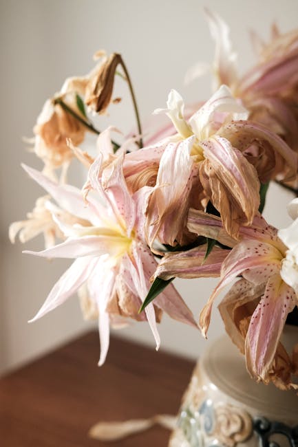 A close-up of a wilted bouquet of pink and cream lilies with speckled petals, showing signs of wilting with browning and curling edges, placed in a vase on a wooden surface. The flowers' petals appear