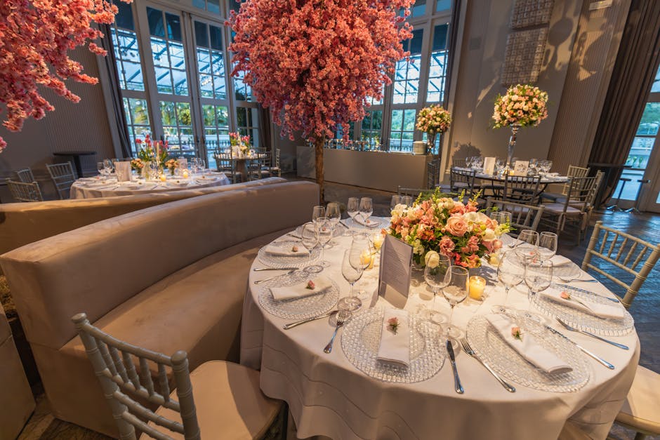 A round banquet table with a white linen tablecloth is elegantly set for a formal event, featuring multiple clear glass wine glasses, silver cutlery, and neatly folded white napkins on transparent, te
