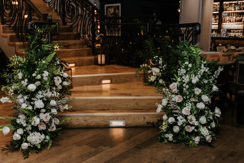 Two large floral arrangements featuring white and pale pink roses, white gerbera daisies, and lush green foliage flank a set of wooden stairs with built-in lighting, inside a sophisticated venue. The 